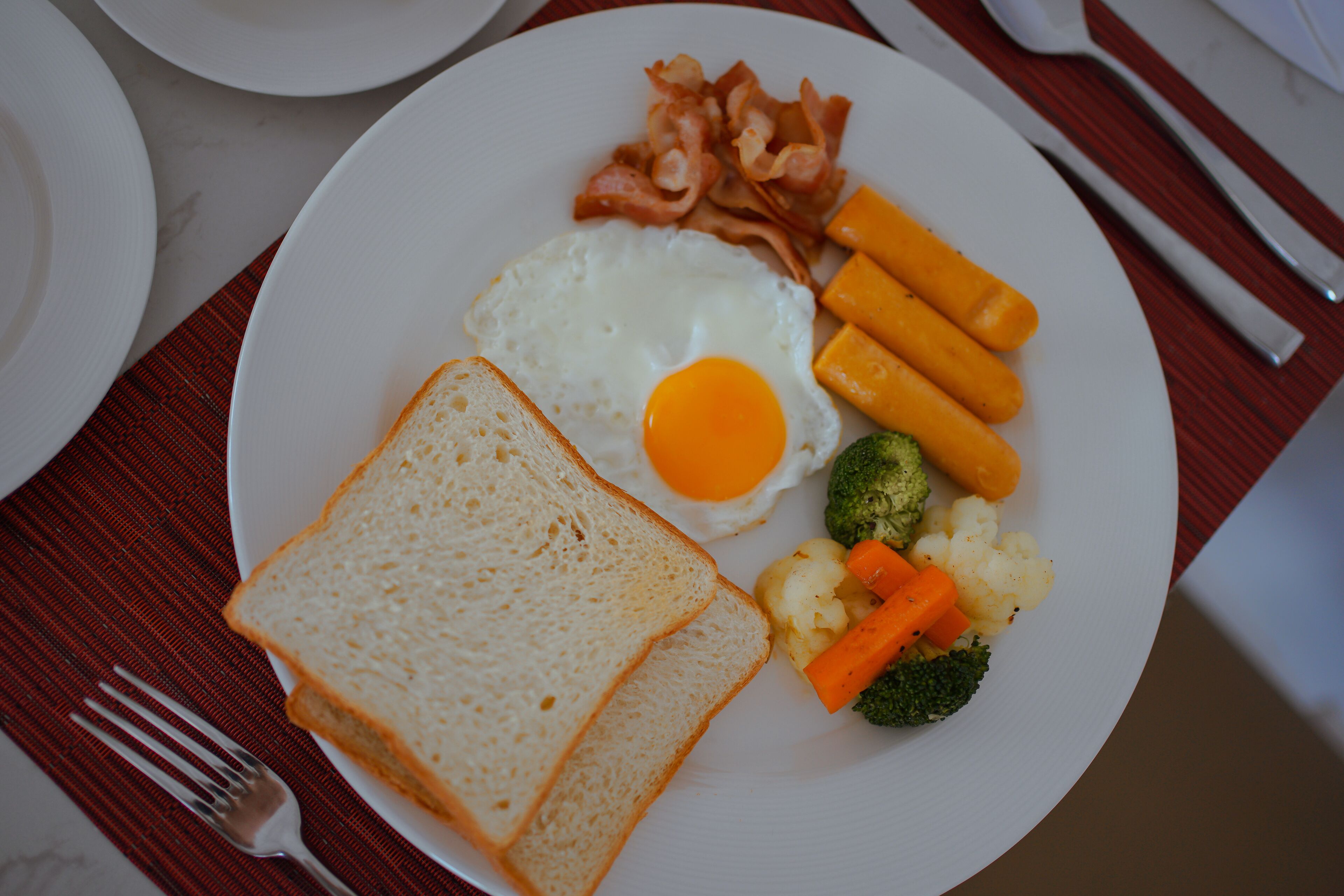 A plate with toast, a fried egg, bacon, carrots, broccoli, and cauliflower.