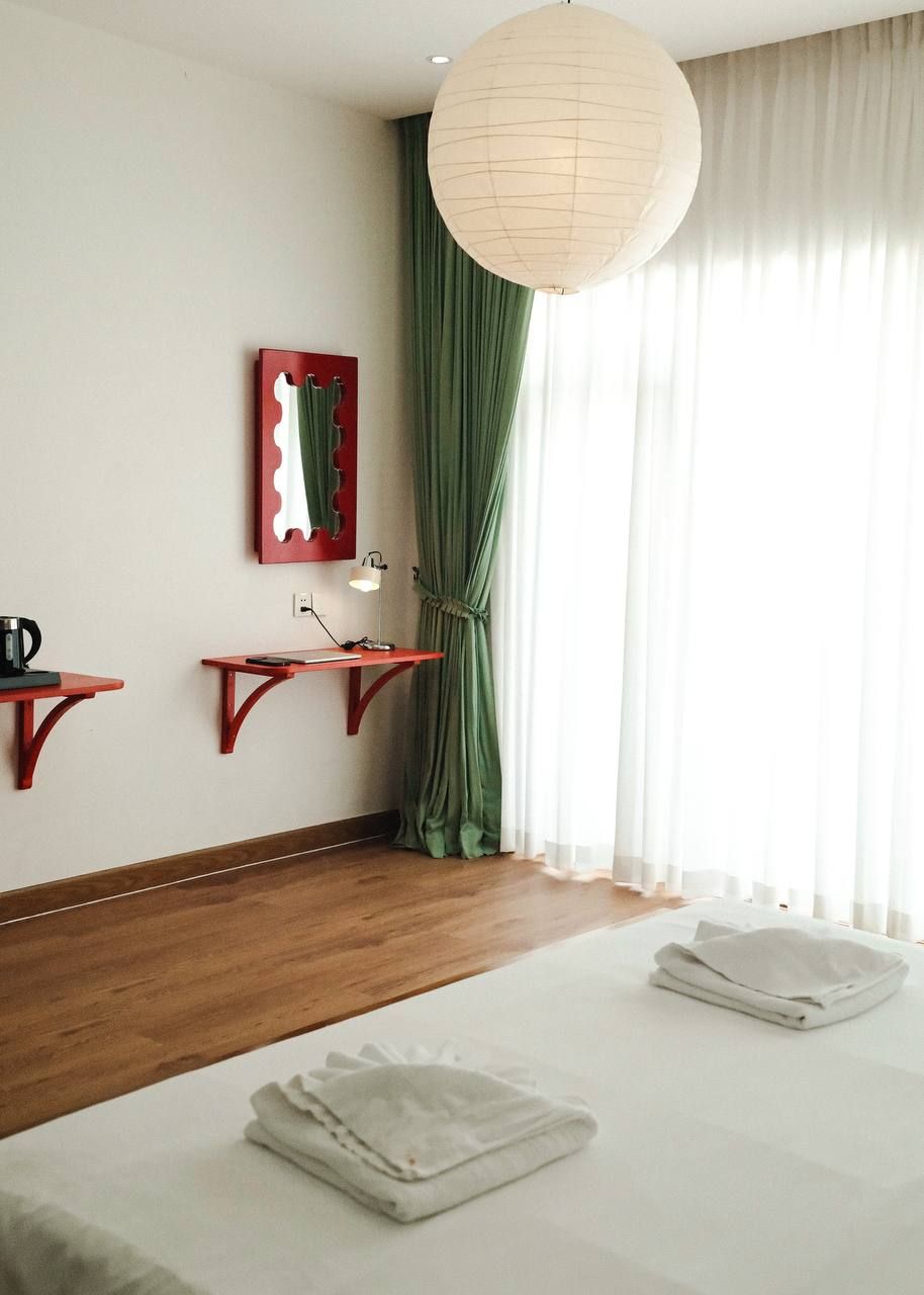 A hotel room with a wooden floor, a bed with white linens, a nightstand with a lamp, a red-framed mirror, and a large window with sheer curtains.