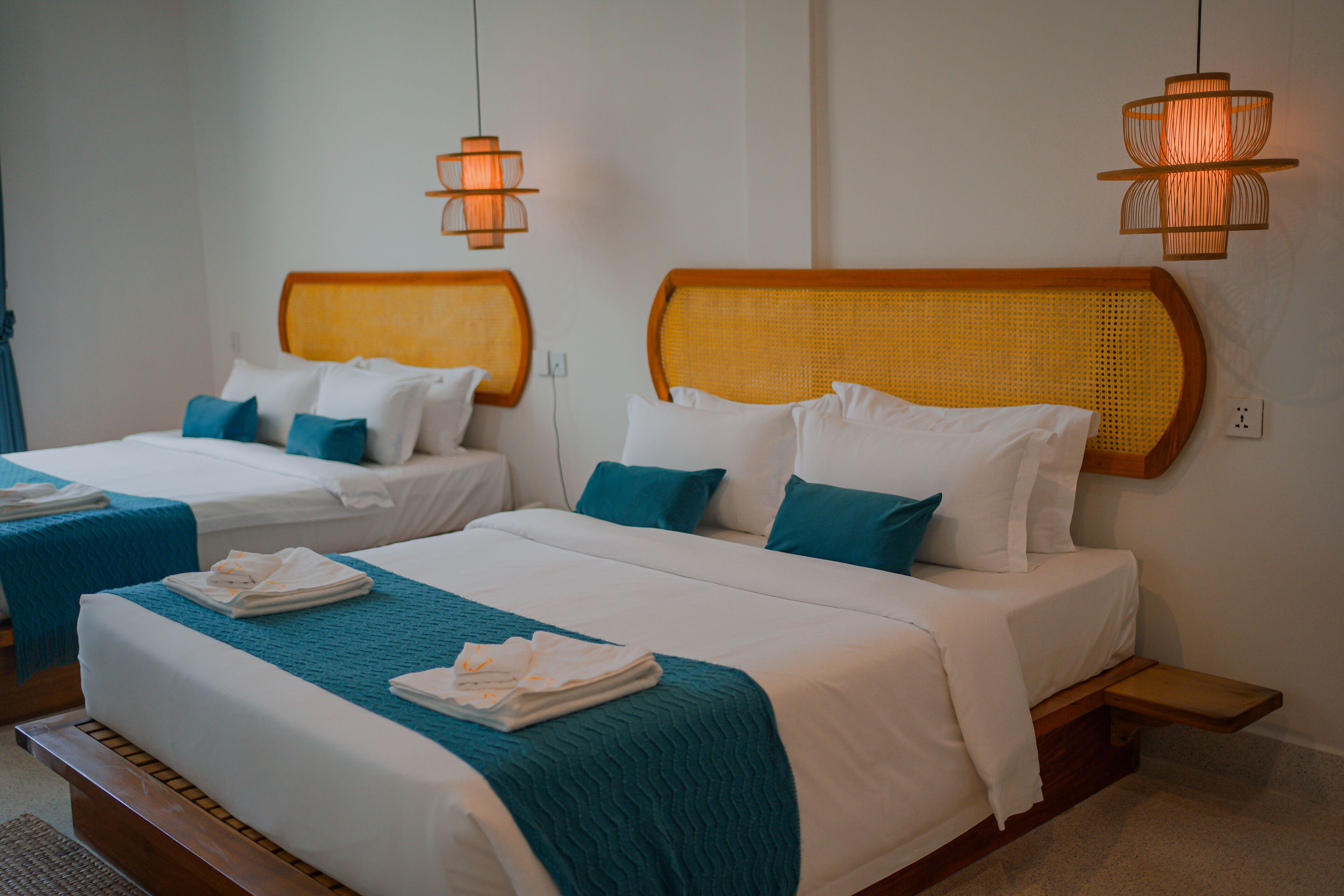 Quarto de hotel com duas camas, cada uma com cabeceira, roupa de cama branca e almofadas decorativas azul-turquesa. Há duas luminárias fixadas na parede e uma pequena estante de madeira.