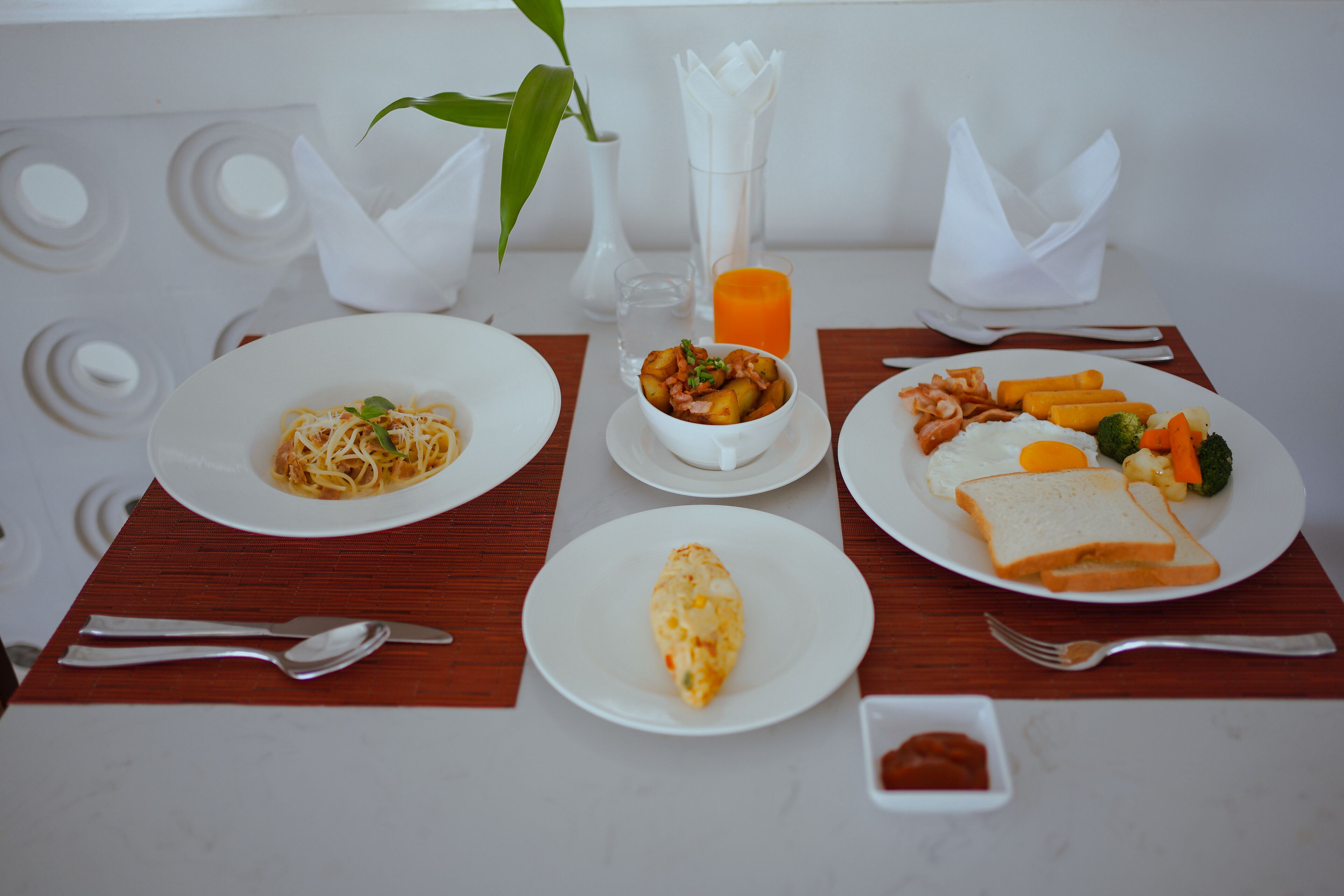 Una mesa de comedor con platos de comida, cubiertos y un vaso de zumo de naranja.