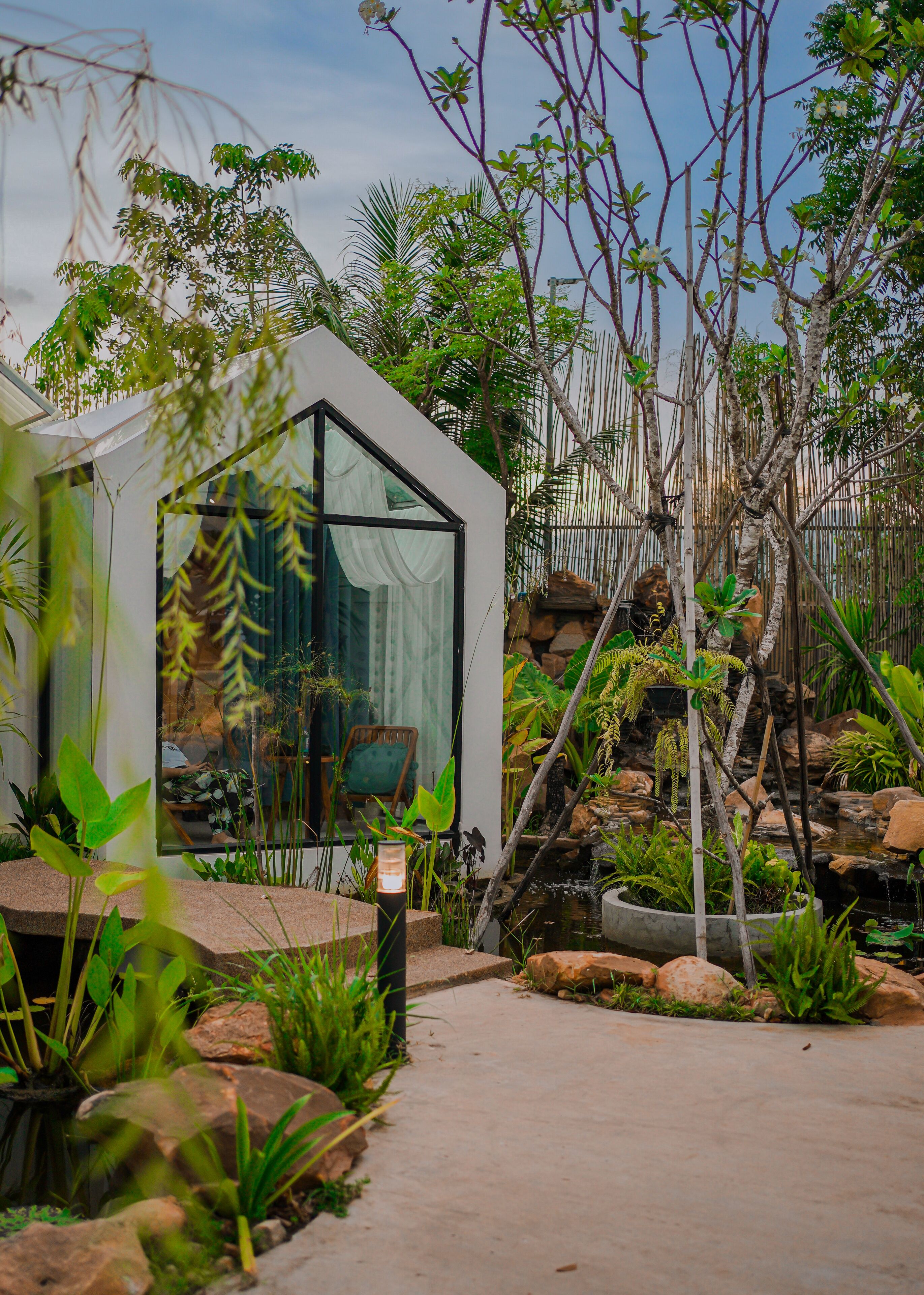Una moderna casa in vetro con vista su un rigoglioso giardino, che include uno stagno e diverse piante.