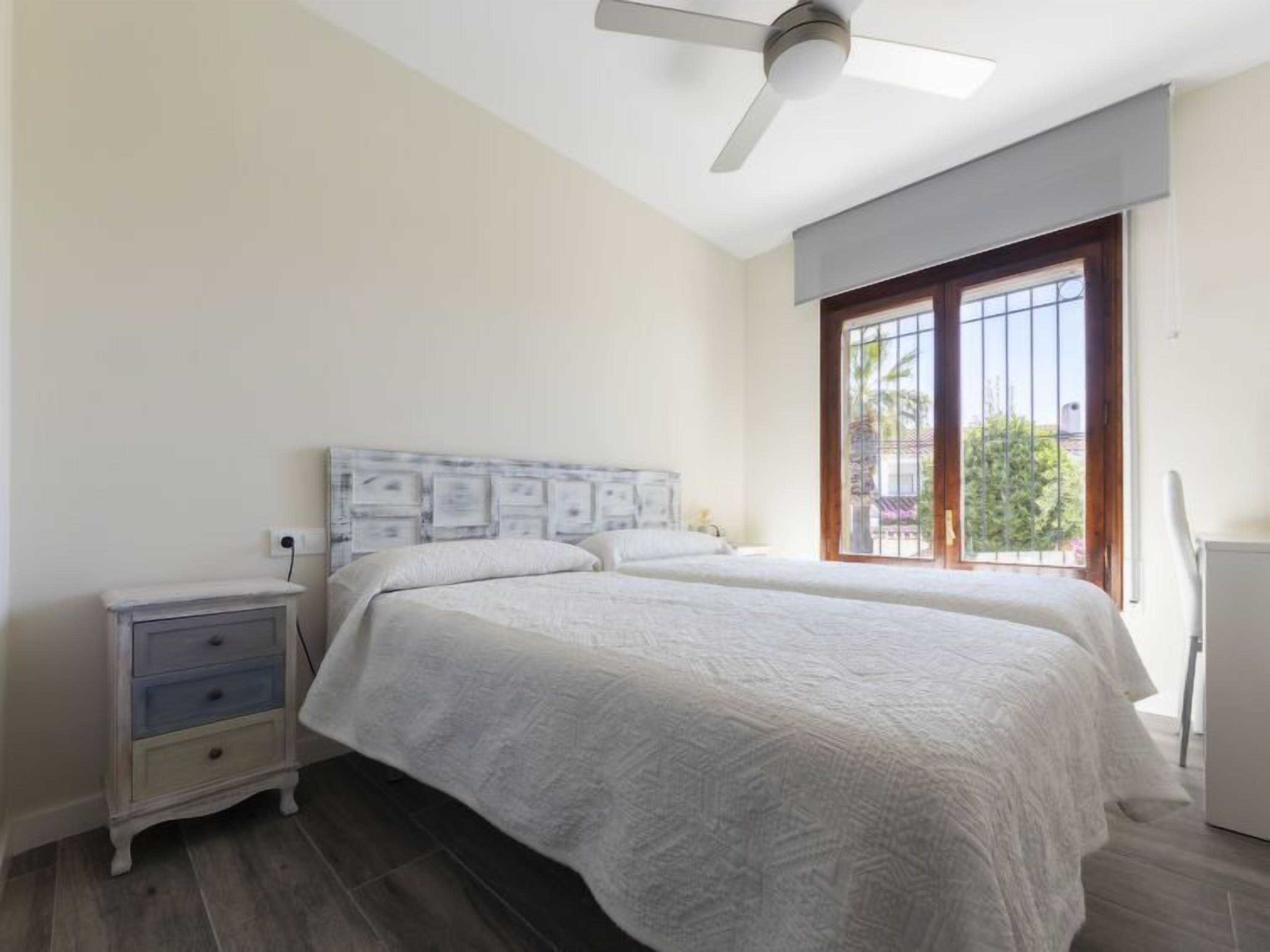 A bedroom with two beds, a ceiling fan, a nightstand, and a window with bars.
