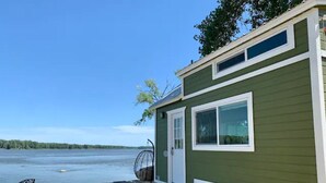 Una casa móvil verde con un pequeño porche, una silla y una alfombra junto a un lago.