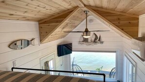 Un dormitorio con cama, ventilador de techo, ventana con vista al agua y televisor montado.