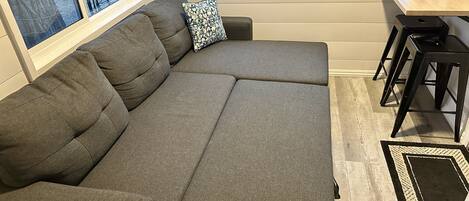 A gray sectional sofa with a patterned pillow, a small table with a framed picture, and a view of a body of water through a window.