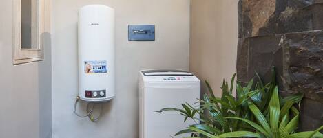 A water heater and a washing machine in a corner with a stone wall and a potted plant.
