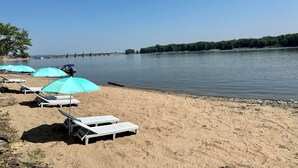 Une plage avec des transats et des parasols, une rivière paisible et un pont au loin.