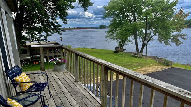 Une terrasse en bois avec deux chaises bleues et un pot de fleurs, donnant sur un lac o ù se trouvent une table de pique-nique et un banc.