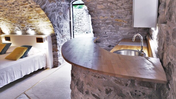 Una habitación con paredes de piedra, una encimera de madera curvada y un lavabo. Se observa una cama con almohadas y una puerta que da a un espacio exterior.