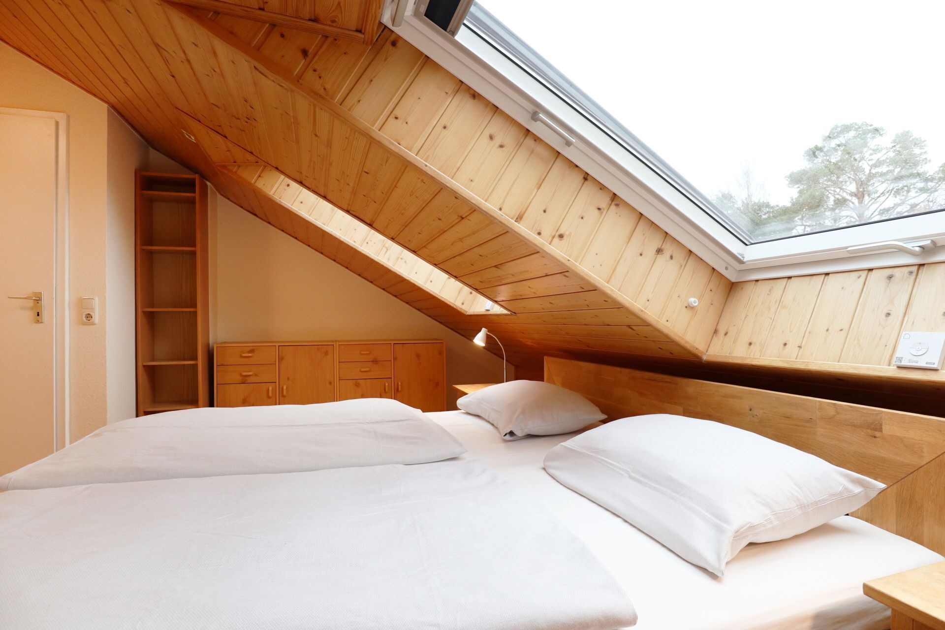A bedroom with a sloped ceiling, a skylight, a wooden wardrobe, and a bed with white bedding.