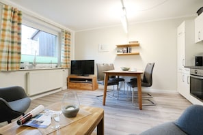 A modern living room with a TV, dining table, and chairs. There is a kitchen area with appliances and a window with curtains.
