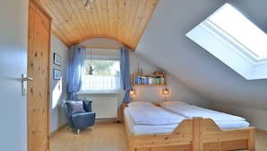 A bedroom with a sloped ceiling, a wooden bed, a chair, a window with curtains, and a shelf with books.