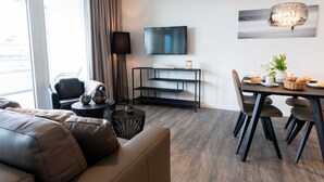 En moderne stue med en lædersofa, et spisebord til fire personer, et fjernsyn ophængt på væggen og et stort vindue med gardiner.
