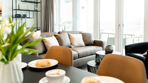 En moderne stue med sofa, sofabord og spisebord. Der er et bogreol og et stort vindue med gardiner.
