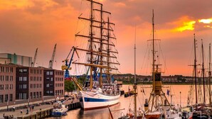 En havn med et stort sejsskib ved kaj, omgivet af andre både og bygninger.