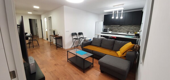 A modern living room with a sofa, coffee table, and a kitchen area with a refrigerator, microwave, and cabinets.