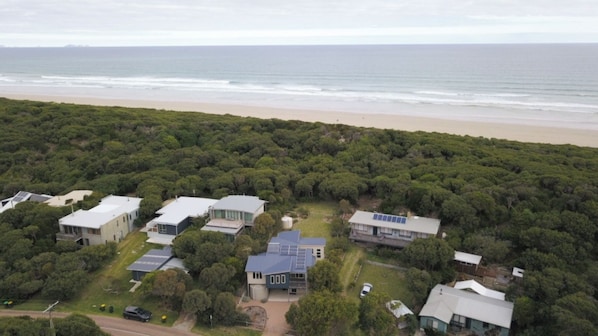 A coastal residential area with houses surrounded by dense greenery, a beach in the background, and solar panels on some buildings.
