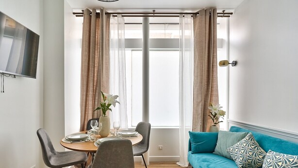 Un coin repas avec une table ronde, deux chaises et un vase rempli de fleurs. On y trouve également un canapé avec des coussins à motifs et un téléviseur fixé au mur.