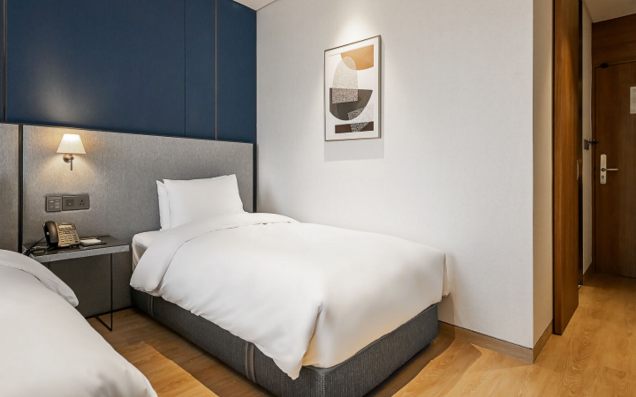 A hotel room with a bed, bedside table, lamp, and a framed abstract artwork on the wall.
