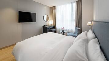 Habitación de hotel con una cama grande, un escritorio con silla, un televisor en la pared y un ventanal con cortinas.