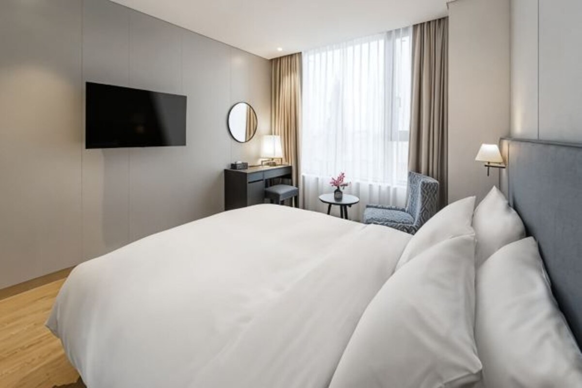 A hotel room with a large bed, a desk with a chair, a TV mounted on the wall, and a window with curtains.