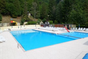 A rectangular swimming pool with a red slide, surrounded by white lounge chairs and umbrellas, set against a backdrop of trees and a hillside. - Inviting 2-bed House in Tignale (Tignale)