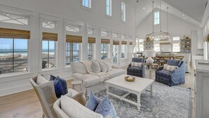 A spacious living room with a large window overlooking the beach, a dining area with a chandelier, and a kitchen with white cabinets.