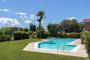 A rectangular swimming pool with a clear blue water, surrounded by a well-maintained lawn, palm trees, and a red-roofed building in the background. - Regarda - renewed holiday apartment "Blitz"  with pool, pet allowed (Lazise)