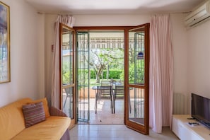 A room with a sofa, a TV, a sliding glass door leading to an outdoor area with a table and chairs, and a sign indicating an outdoor area.