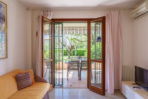 A room with a sofa, a TV, a sliding glass door leading to an outdoor area with a table and chairs, and a sign indicating an outdoor area.