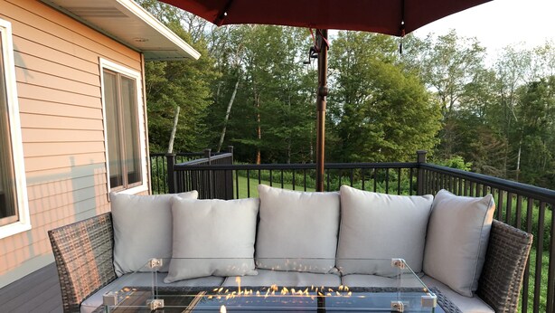 A glass-top fire pit with burning logs, surrounded by a cushioned outdoor seating area with a view of a wooden house and trees. - Mountain View Getaway (West Cornwall)