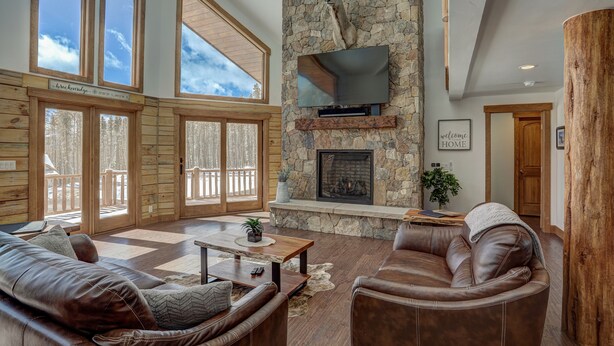 A spacious living room with a stone fireplace, leather sofas, and large windows offering a view of the outdoors. - Modern Luxury Breckenridge Cabin | Hot Tub (Breckenridge)