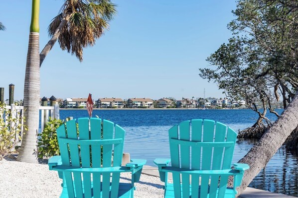 Two turquoise Adirondack chairs facing a waterfront with a view of houses and a palm tree.