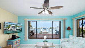Un salon comprenant un téléviseur, un ventilateur de plafond, une grande fenêtre donnant sur un quai et des bateaux, une petite table avec une plante et un canapé.