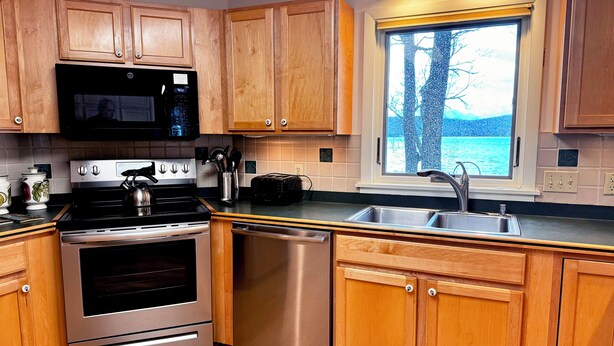 A modern kitchen with wooden cabinets, stainless steel appliances, a window with a view of trees, and a sink. - Large house on beautiful lake, adjoins Sleeping Bear Dunes National Lakeshore (Empire)