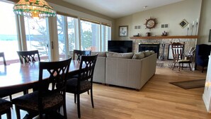 A spacious living room with a wooden floor, a sofa, a dining table with chairs, a TV, and a fireplace. - Large house on beautiful lake, adjoins Sleeping Bear Dunes National Lakeshore (Empire)