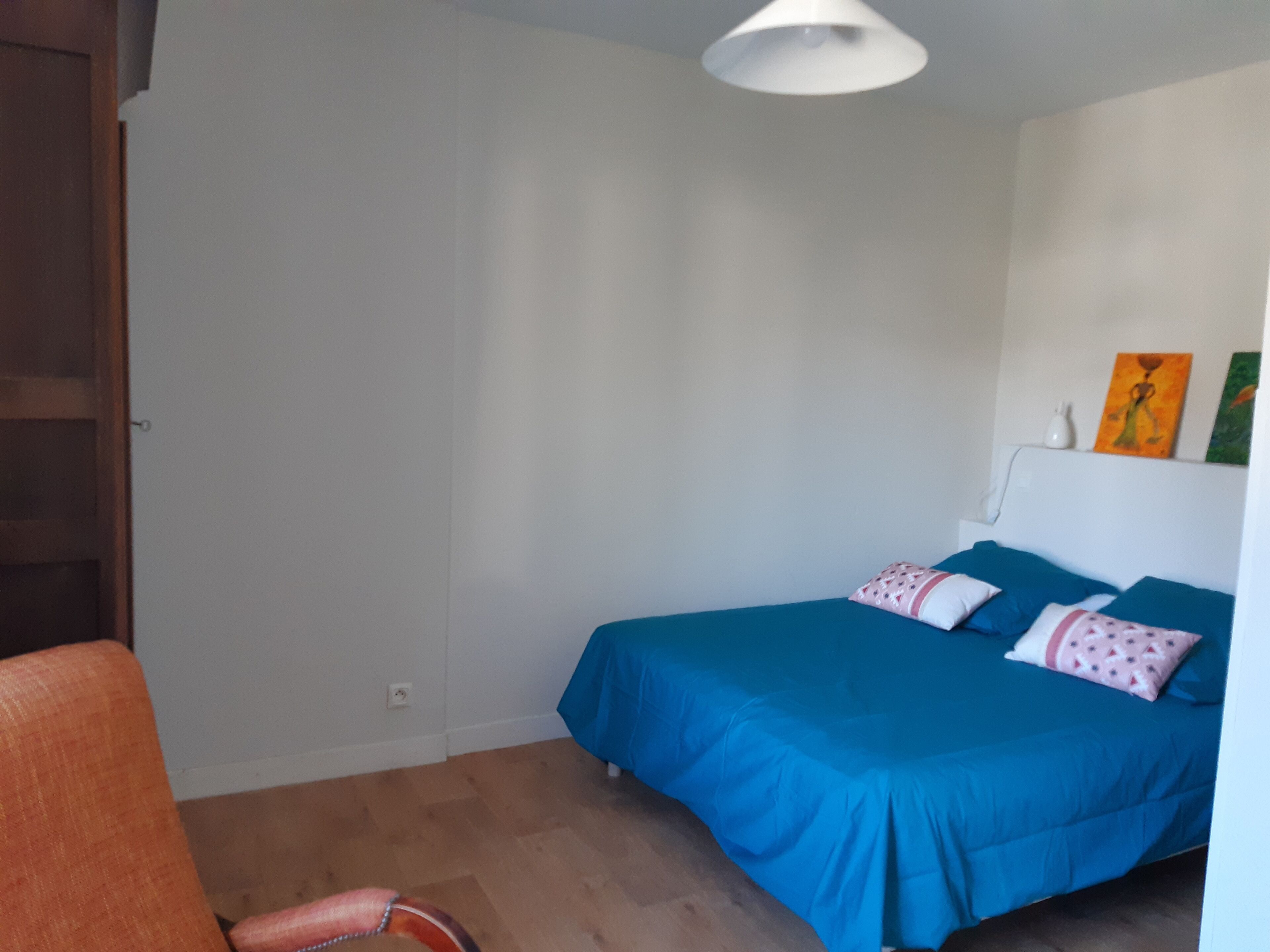 Un dormitorio con cama, una silla, una lámpara de techo blanca y una pared con dos cuadros.