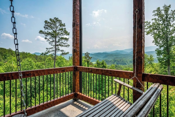 A wooden deck with a swing, surrounded by metal railings, overlooking a forested landscape.