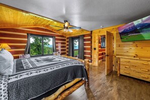 A log cabin bedroom with a bed, a ceiling fan, a window, and a view of the outdoors.