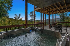 A hot tub with a wooden deck and chairs, overlooking a mountainous landscape.