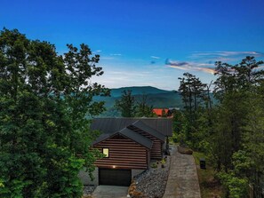 A modern log cabin with a garage, surrounded by dense forest and a mountain backdrop.