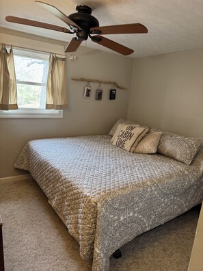 Quarto com cama, ventilador de teto, janela com cortinas e prateleira na parede com quadros.