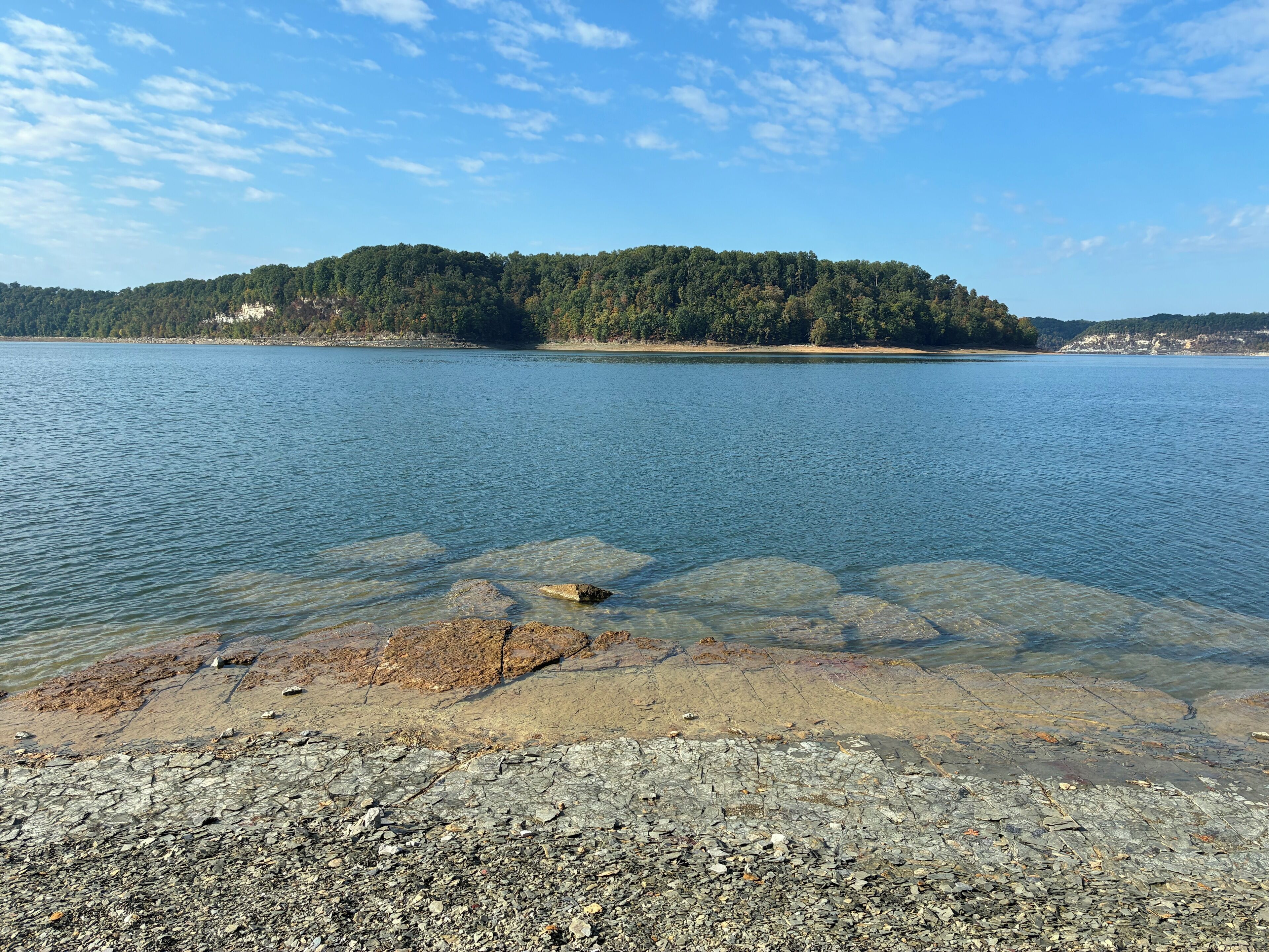 A lake with clear water, a rocky shoreline, and a forested hill in the background.