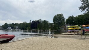 Una playa arenosa con un muelle que se adentra en un lago, rodeado de árboles y casas.