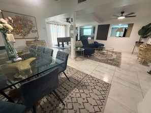 A spacious living area with a glass dining table, comfortable seating, a ceiling fan, and a view into the kitchen.