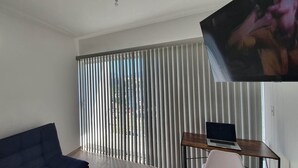 Un salon moderne avec un canapé bleu, une petite table en bois sur laquelle se trouve un ordinateur portable, et une grande fenêtre équipée de stores verticaux.