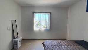 Une chambre à coucher avec un lit, une fenêtre avec des stores, un panier à linge et un miroir fixé au mur.