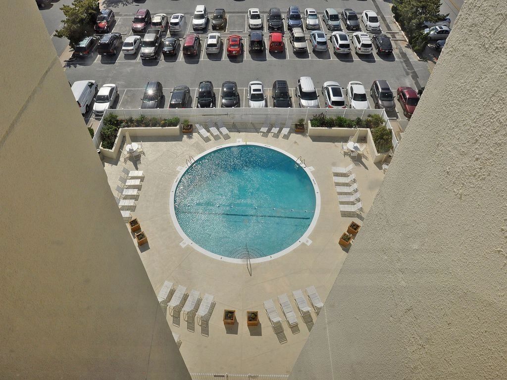 Une vue aérienne d’un hôtel avec une piscine circulaire entourée de chaises longues et un parking rempli de voitures.