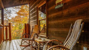 Uma cabana de madeira com varanda, com cadeiras de balanço e uma mesa, um letreiro com a inscrição "BLES AMERICA" e vista da folhagem outonal.