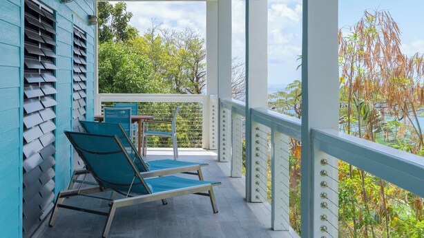 Un espace extérieur aménagé, abrité et doté de deux fauteuils et d’une table, qui offre une vue sur un jardin où poussent différentes plantes et arbres.